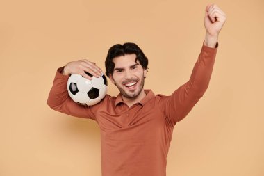 Genç, yakışıklı bir adam sevinçle elini kaldırır futbol topunu tutarken, heyecanını ifade eder..