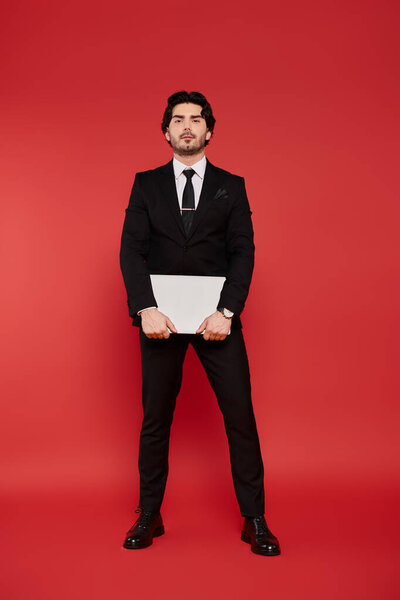 With determination in his eyes, a young man stands strong in a suit, clutching a laptop.