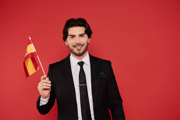 Handsome young man dressed in a suit joyfully waves a small flag symbolizing Spain.