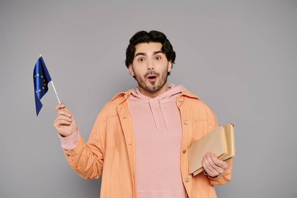 Delightful young man in casual clothing shows enthusiasm while holding a small flag and a book.