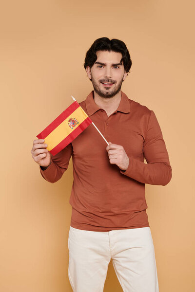Молодой красивый мужчина с гордостью держит маленький испанский флаг, улыбаясь, воплощая культурную гордость.