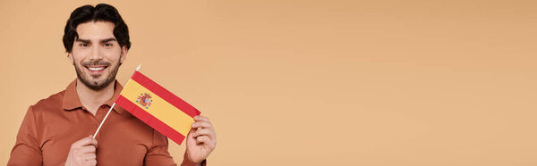 Брюнетка улыбается с маленьким испанским флагом на тёплом, нейтральном фоне.