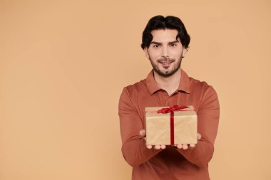 Genç yakışıklı bir adam, güzelce paketlenmiş kırmızı kurdeleyle süslenmiş bir hediyeyi sunarken gülümsüyor..