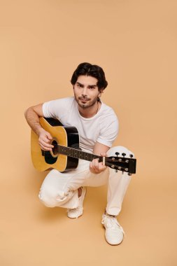 Yakışıklı genç adam gitar çalarken çömeliyor, yaratıcılık ve neşe saçıyor sıcak bir ortamda.
