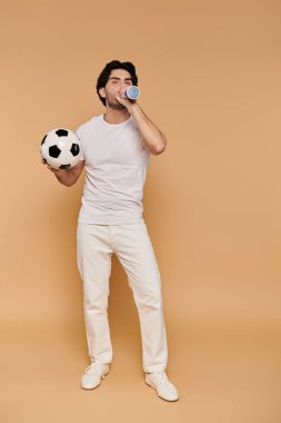 Siyah saçlı genç bir adam futbol topunu tutuyor ve enerji ve neşe saçan bir şişeden içiyor..