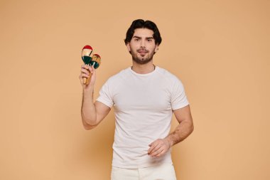 Kumral saçlı genç yakışıklı bir adam açık renkli bir stüdyoda mutlu bir şekilde marakas sallıyor..