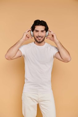 Siyah saçlı genç bir adam kulaklıkla müzik dinliyor, sıcak bir arka planda neşe saçıyor..