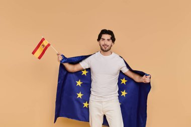 Yakışıklı genç adam Avrupa Birliği bayrağına sarılmış İspanyol bayrağını tutuyor, neşe saçıyor.