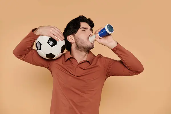 Handsome young man sips a beverage while holding a soccer ball, showcasing a sporty vibe.
