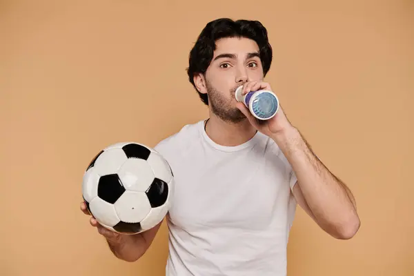 A young handsome brunette man drinks from a bottle while holding a soccer ball, smiling joyfully.