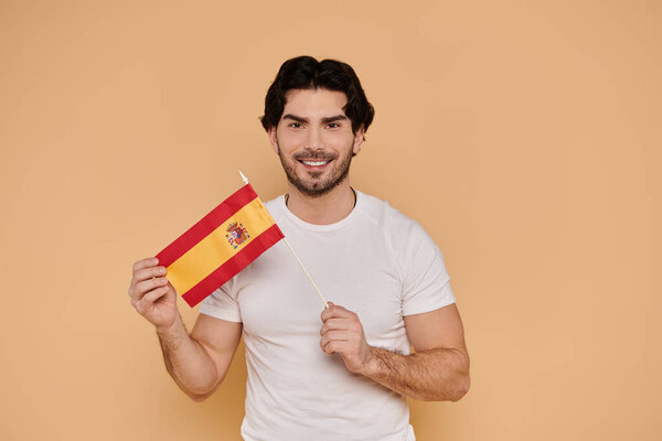Красивый молодой человек улыбается, показывая маленький испанский флаг, излучая радость и гордость.