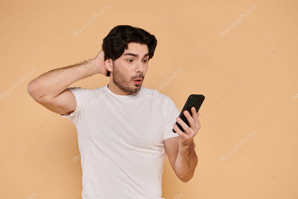 Man with brunette hair stands against an orange background, showing surprise while looking at phone.