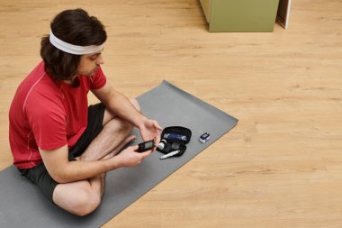 Genç adam, şık dairesinde yoga minderinin üzerinde otururken kan şekerini ölçüyor..