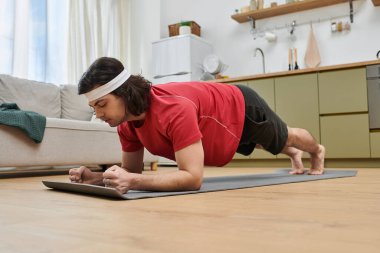 Yakışıklı genç adam, şık dairesinde bir fitness paspası üzerinde tahta egzersizi yapıyor..