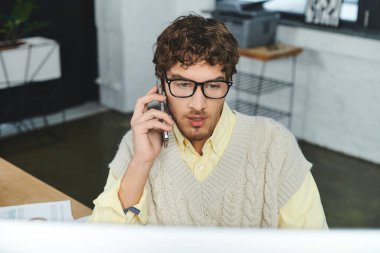 Süveter giyen genç bir adam, parlak bir ofiste masasında çalışırken telefonda konuşuyor..