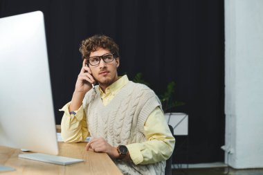 Süveter giyen genç bir işadamı masasında çalışırken telefonla konuşuyor..