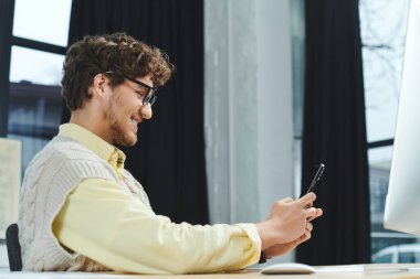 A young man with curly hair smiles while checking his phone in a stylish office environment. clipart