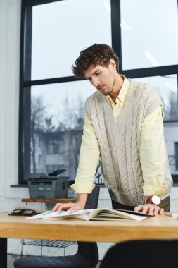Kıvırcık saçlı genç bir adam şık bir ofiste masa başında dururken görevlerine odaklanır..