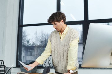 Süveter giyen genç bir adam modern bir şirket ofisinde çalışırken dokümanları dikkatle inceliyor..