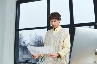 Süveter giyen odaklanmış genç bir adam modern bir ofisteki belgeleri inceliyor..