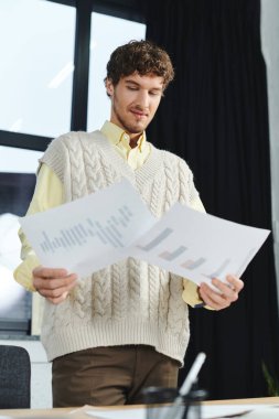 Curly-haired man in a sweater vest studies important paperwork, deep in thought. clipart