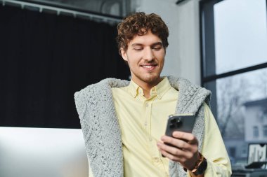 Çağdaş ofis ortamında akıllı telefonunu kontrol ederken kıvırcık saçlı genç bir adam gülümsüyor..