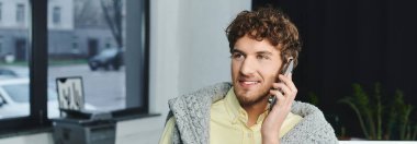 Kıvırcık saçlı genç adam telefonda konuşuyor. İş yerinde şık giyinmiş, afiş yapıyor.