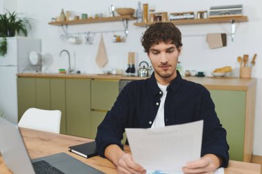 Kıvırcık saçlı bir adam şık bir dairede ahşap bir masada oturur ve önemli evrakları okur..