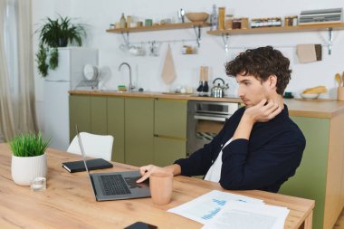 Gömlekli yakışıklı bir genç, modern dekorun ortasında evinde dizüstü bilgisayarına odaklanıyor..