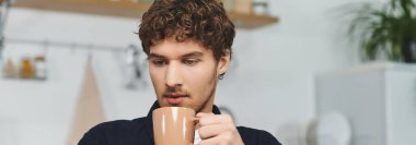 Curly-haired young man sits in his contemporary apartment, savoring a delicious drink, banner clipart