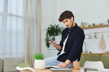 Yakışıklı genç adam şık dairesinde rahat bir şekilde oturuyor, akıllı telefonuna odaklanmış..