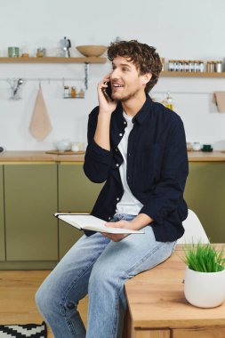 Kıvırcık saçlı bir adam modern bir dairede oturuyor, telefonla konuşurken gülümsüyor..