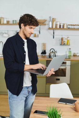 Kıvırcık saçlı yakışıklı bir adam modern bir apartman dairesinde dizüstü bilgisayarına odaklanıyor..