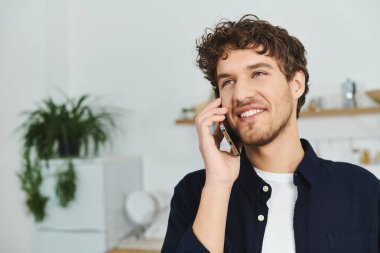 Kıvırcık saçlı genç adam şık evinde telefonla konuşurken gülümsüyor..