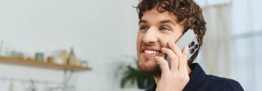 Şık tişörtlü kıvırcık saçlı bir adam çağdaş yaşam alanında akıllı telefondan mutlu bir şekilde sohbet ediyor.