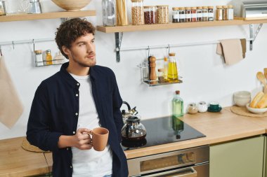Yakışıklı bir genç adam modern mutfağında duruyor, kahvesini yudumluyor ve evinin tadını çıkarıyor..