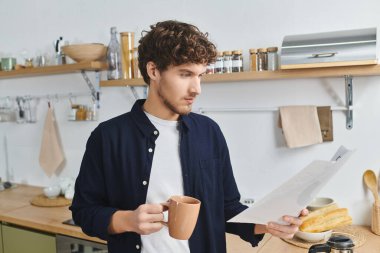 Modern bir mutfakta kıvırcık saçlı bir adam elinde sıcak bir bardak tutarken bir belge okur..