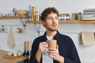 Yakışıklı genç adam, modern mutfak alanını yansıtırken elinde bir bardak tutuyor..