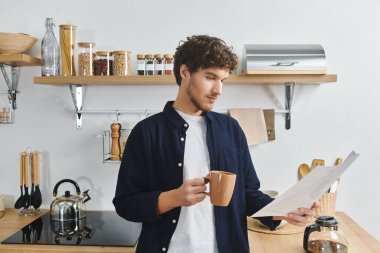 Yakışıklı genç bir adam, şık dairesinde kahvesini yudumlarken bir belge okuyor..