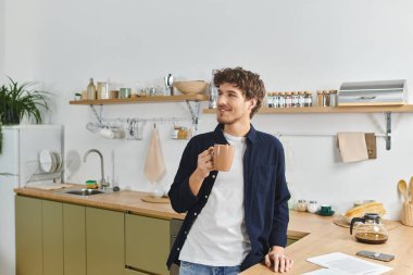 Genç kıvırcık saçlı bir adam şık mutfağında kahvesini yudumlarken rahatlıyor..