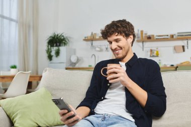 Genç adam şık dairesinde rahatlar, kahvesini yudumlar ve akıllı telefonuyla meşgul olur..