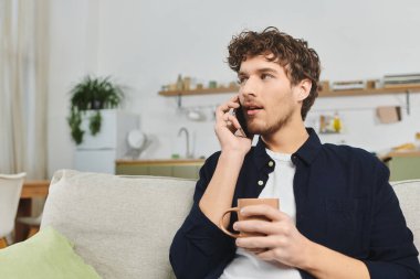 Handsome young man with curly hair relaxes at home, holding a cup and chatting on the phone. clipart