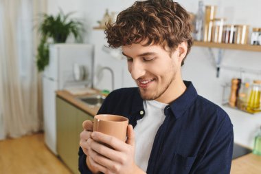 Gömlekli kıvırcık saçlı adam evde sıcak bir içecek tutarken mutlu bir şekilde gülümsüyor..