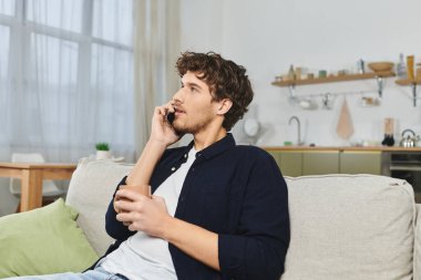 Kıvırcık saçlı genç adam bir telefon görüşmesi yapıyor, evinde rahatça oturuyor..