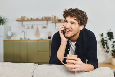 Kıvırcık saçlı neşeli genç bir adam telefonda konuşurken elinde bir bardak tutuyor..
