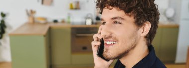 Şık bir mutfak ortamında telefonuyla konuşan kıvırcık saçlı genç bir adam gülümsüyor.