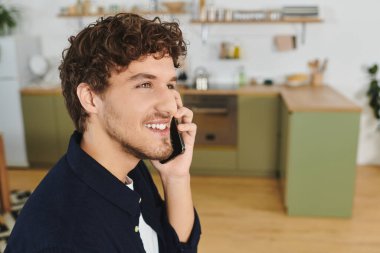 Kıvırcık saçlı genç bir adam şık mutfağında telefonla konuşurken gülümsüyor..