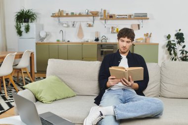 Kıvırcık saçlı yakışıklı genç bir adam parlak dairesinde kitap okurken rahatlıyor..