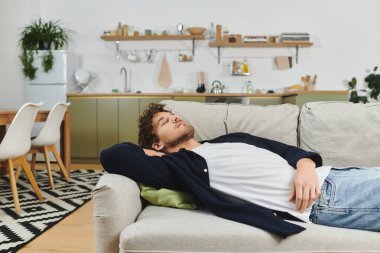 A young man with curly hair enjoys a peaceful moment lying on the sofa in a modern apartment. clipart