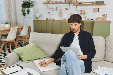 A young man sits comfortably in his modern apartment, jotting down notes while deep in thought. clipart